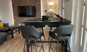 A modern dining room with a black table and high chairs. In the background, there is a television and a decorative stone wall.
