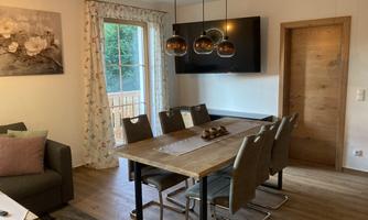 A modern dining area with a large wooden table and comfortable chairs. A TV hangs on the wall, and large windows provide plenty of natural light.
