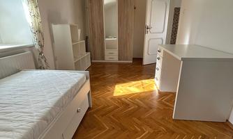 A bright room with a single bed, several pieces of furniture, and a wardrobe. The floor is wooden, and there is plenty of natural light.