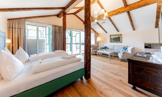 A bright room with a wooden beam ceiling and large windows. It contains a double bed and a cozy seating area.