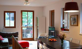 A modern living room with large windows and a bright, welcoming atmosphere. The space is furnished with a dining area, a sofa, and decorative plants.