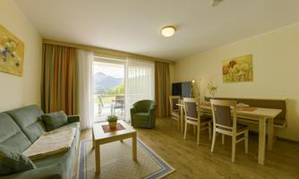 A bright and cozy living room setup with a sofa, a dining area, and access to the balcony. Large windows offer a view of the mountains.