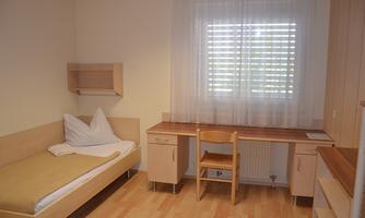 A simple room with a single bed, a desk, and a window with blinds. The walls are light and the floor is made of wood.