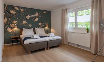A cozy bedroom with a large double bed and a decorative wall pattern. The room is bright and inviting with a window view to the outside.