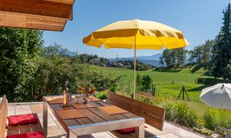 An inviting outdoor area with a wooden table and chairs under a yellow sun umbrella. The view shows a green landscape with trees and mountains in the background.