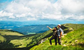 Two hikers are standing on a hill and pointing at the view. In the background, green meadows and mountains can be seen under a clear sky.