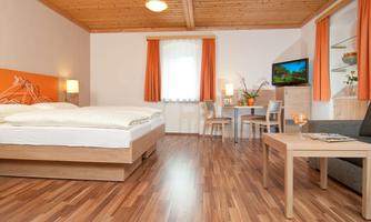 A cozy room with a double bed, a small table, and a sofa. The walls are decorated in warm colors and the lighting is pleasant.
