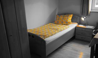 A simple bedroom with a single bed and a yellow checked duvet cover. Next to the bed is a bedside table with a lamp.