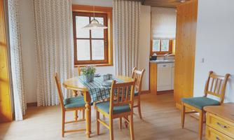 A cozy dining area with a round table and chairs. The windows let in plenty of daylight, and the kitchen is visible in the background.