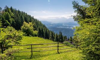 A picturesque view of green meadows and forests under a clear blue sky. In the background, mountains and a beautiful view into the valley can be seen.