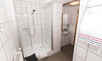 A modern bathroom with a walk-in shower and white tiles. There is a sink and a small shelf with care products.