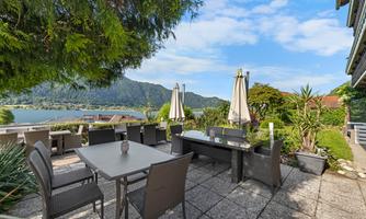 A cozy terrace with a table and chairs. In the background, the lake and the mountains are visible, surrounded by plants.