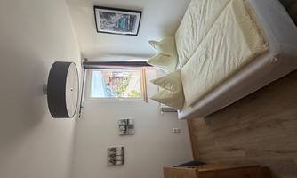 A cozy bedroom with a double bed and beige bed covers. The room has a television, a wooden wardrobe, and plenty of daylight through the window.
