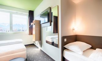 A modern hotel room with a bed, a small sleeping area, and a large window that offers a view of the mountains. The walls are painted in soft colors and a large mirror wall reflects the brightness of the room.