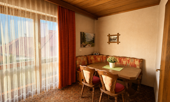 A cozy dining area with a wooden chair and table. Curtains hang in front of the window front, and there is a nice view outside.