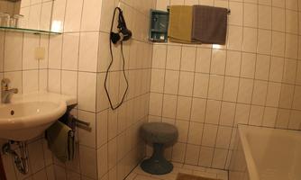 A small bathroom with white tiles. There is a sink, a bathtub, and a stool.
