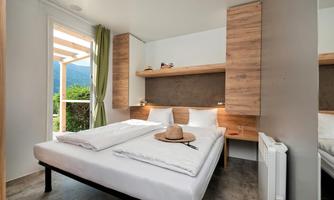 A bright room with a double bed and modern wooden interior. Large windows offer a view of greenery.