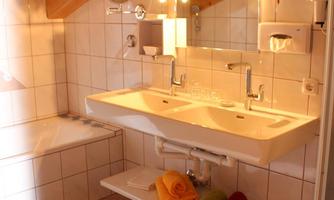 A modern bathroom with a wooden ceiling and bright tiled floor. There is a double sink, a bathtub, and colorful towels on a shelf.