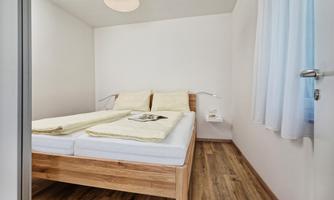 A bright bedroom with a double bed and wooden floor. The walls are white, and there is a window with curtains.