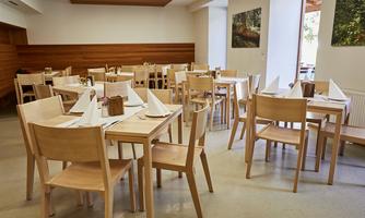A modern dining room with wooden chairs and tables. The tables are set for guests and there are large windows that let in plenty of light.