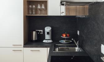 A modern kitchen with white and wooden cabinets. On the countertop, there is a coffee machine, a kettle, and a plate with apples.