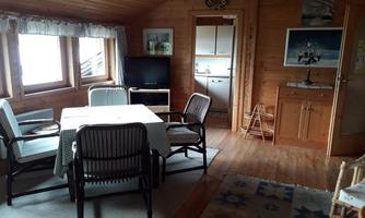 A cozy living space with wood paneling, a dining table and chairs. Windows with a view outside and a small kitchenette are also visible.