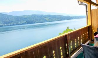A beautiful view of a calm lake surrounded by mountains. The sight is characterized by a terrace with a wooden lattice and a chair.
