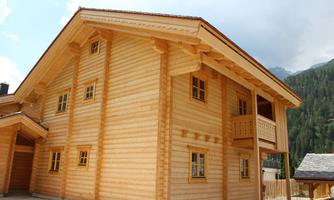 A modern wooden house with many windows and a balcony. In the background, green mountains can be seen.
