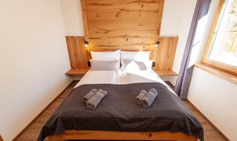 A cozy bedroom with a large bed and wooden details. There are bright walls and windows that let in daylight.