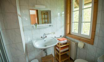 A small bathroom with white tiles and a sink. In the background, towels are placed on a shelf next to a window looking outside.