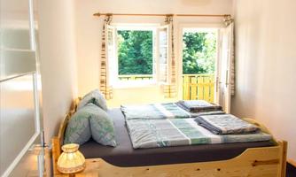 A bright bedroom with a double bed and two windows. Green trees are visible through the windows.