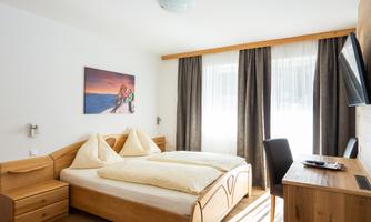 A cozy bedroom with a double bed and wooden furniture. Large windows let in plenty of daylight, and a wall mural provides a nice accent.