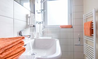A modern bathroom with a sink and a shower.  
Orange towels are neatly stacked.