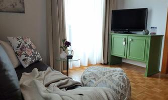 A cozy living room with a beige sofa and a green TV unit. The room is brightly lit with a large window and curtains.