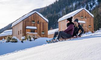 Two women are tobogganing on a snowy slope. In the background, there are modern wooden-clad huts and a beautifully snow-covered winter landscape.