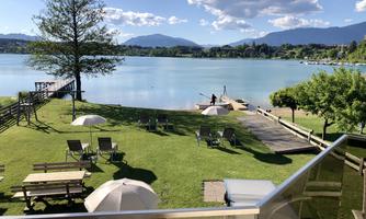An idyllic landscape by the lake with a green meadow, loungers, and sun umbrellas. In the background, the mountains and a clear blue sky are visible.