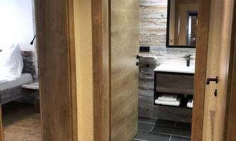 A modern hallway with two doors. On the left side is a bedroom and on the right side is a bathroom featuring wood and tile design.