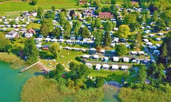 A spacious campground landscape by the water with numerous trailers and tents. Surrounded by trees and green spaces, the site offers a relaxed atmosphere.