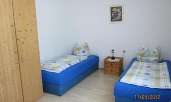 A simple bedroom with two blue beds and light furniture. A picture hangs on the wall, and a nightstand stands between the beds.