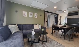 A modern living space with a comfortable, dark gray couch and a dining table. The room is bright and invitingly designed, with plants and artwork on the walls.