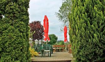 A well-maintained path leads to a cozy outdoor area with green chairs and red umbrellas. Tall cypress trees and flower frames surround the terrace.