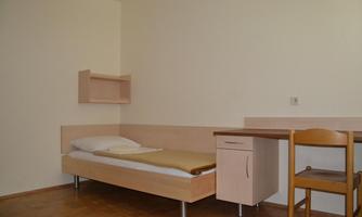 A simple room with a bed, a table, and a chair. The walls are light and the floor is made of wood.