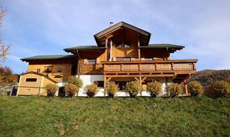A charming wooden house with a large balcony and a green meadow in front of it. The surroundings are hilly and showcase a picturesque landscape.