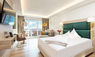 A modern hotel room with a large bed and a cozy seating area. Large windows offer a view of a balcony and plenty of natural light.
