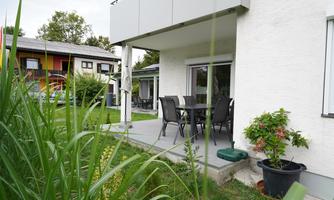 A terrace with a dining table and chairs, surrounded by green grass and plants. In the background, additional houses are visible.