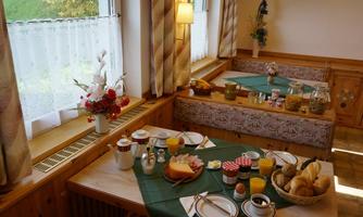 A cozy breakfast table with various foods is set in a bright room. The table is arranged with rolls, cheese, cold cuts, fruit, and drinks.