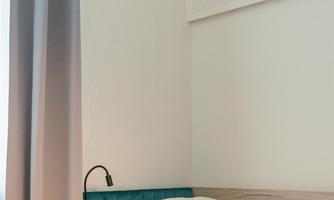A simple bedroom with a bed, a nightstand, and a lamp. The walls are light, and the window is covered with a curtain.