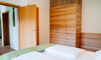 A modern bedroom with a double bed and a wooden wardrobe. The walls are bright, and there is an open door leading to another room.