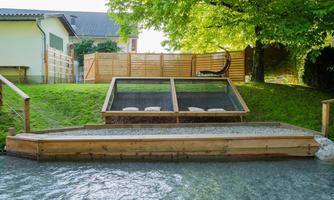 A beautiful garden with a wooden walkway and a small pond. In the background, there is a seating area and a green tree canopy visible.