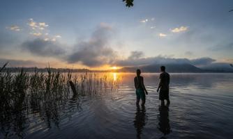 A sunset over a calm lake with two people in the water. In the foreground, there are tall grasses and the soft light reflects off the water's surface.